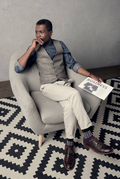 stylish african american man in waistcoat sitting in armchair with business newspaper - Фото, изображение
