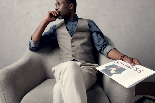 вдумчивый африканский американец сидит в кресле с деловой газетой
 - Фото, изображение