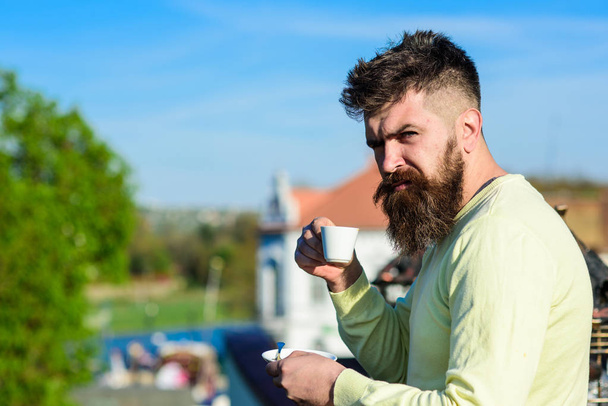 Bearded man with espresso mug, drinks coffee. Man with beard and mustache on strict face drinks coffee, urban background, defocused. Coffee break concept. Man with long beard looks strict and serious - Фото, зображення