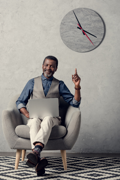 stijlvolle Afro-Amerikaanse zakenman die omhoog wijst en laptop in office gebruikt met klok op muur - Foto, afbeelding