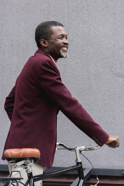elegante hombre afroamericano sonriente en chaqueta borgoña posando cerca de la bicicleta
 - Foto, Imagen