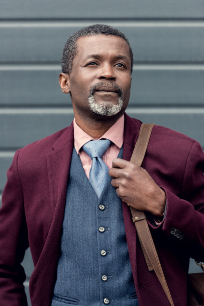 stylish thoughtful african american man in burgundy jacket with bag - Fotoğraf, Görsel