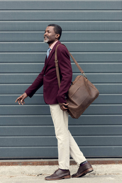 stylish mature african american man posing in trendy burgundy jacket with leather bag - Фото, зображення