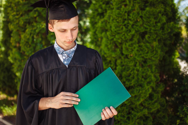 Επιτυχημένη απόφοιτος τύπος, στο ακαδημαϊκό φορέματα, στο αποφοίτηση θέτοντας, κρατώντας ένα πράσινο φάκελο και κοιτώντας το, μελέτη αποχαιρετιστήριο, μπορούν να χρησιμοποιηθούν για τη διαφήμιση, - Φωτογραφία, εικόνα