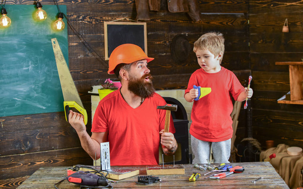Father, parent with beard in protective helmet teaching little son to use different tools in school workshop. Boy, child cheerful playing and learning to use tools with dad. Fatherhood concept - Фото, изображение