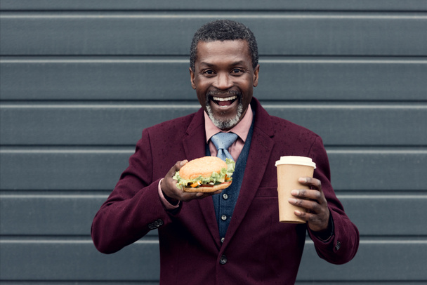 podekscytowany stylowy mężczyzna african american z kawa na wynos i hamburger - Zdjęcie, obraz