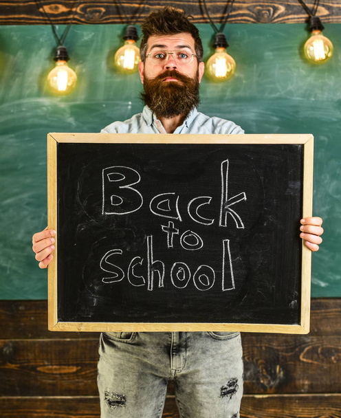 High school concept. Teacher in eyeglasses holds blackboard with title back to school. Man with beard and mustache on calm face welcomes students, chalkboard on background - Foto, imagen