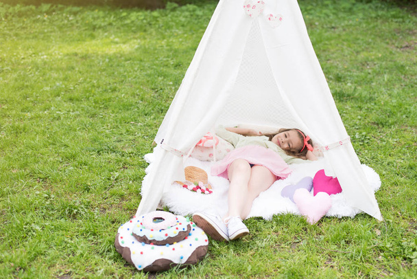 Pieni tyttö makaa ja leikkii teltassa, lasten talon wigwam puistossa. Hyvää äitienpäivää. Lapsi, jolla on isot donitsit, olkihattu ja sydämen muotoiset tyynyt. Kesä, onnellinen lapsuus käsite. Söpö tunteita tyttö vaaleanpunainen keula
. - Valokuva, kuva