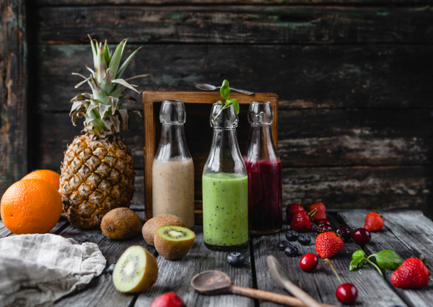 Органические фруктовые коктейли в стеклянных бутылках на деревянном фоне с разнообразными фруктами
 - Фото, изображение
