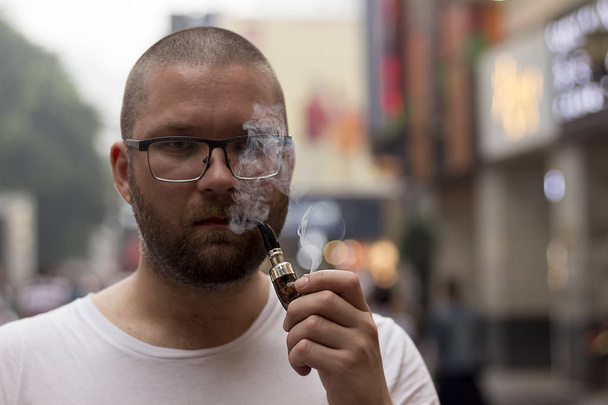 White caucasian handsome bearded man and stylish glasses smoking pipe on the street. Close up portrait. - Photo, Image