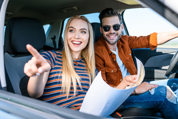 young woman with map in hand pointing by finger to smiling boyfriend sitting behind car wheel - Fotoğraf, Görsel