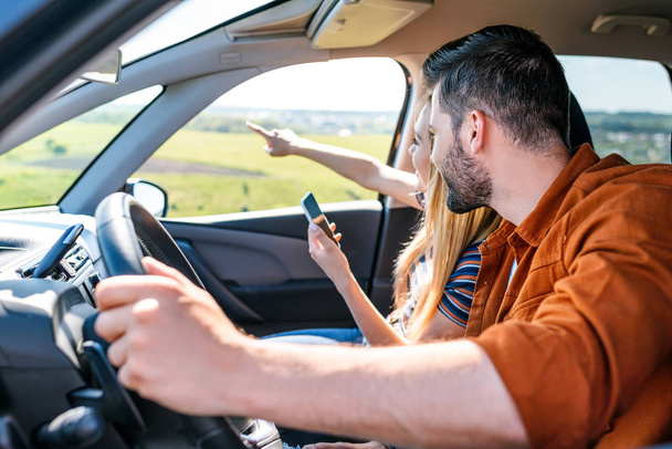 vrouw met smartphone in de hand te wijzen door vinger vriendje zitten achter de auto wiel  - Foto, afbeelding