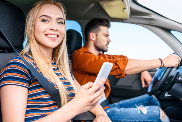 smiling young woman holding smartphone in hand while her boyfriend driving car  - Foto, afbeelding