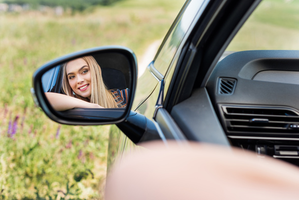 close up view of smiling young woman sitting in car and looking at side mirror  - Fotografie, Obrázek