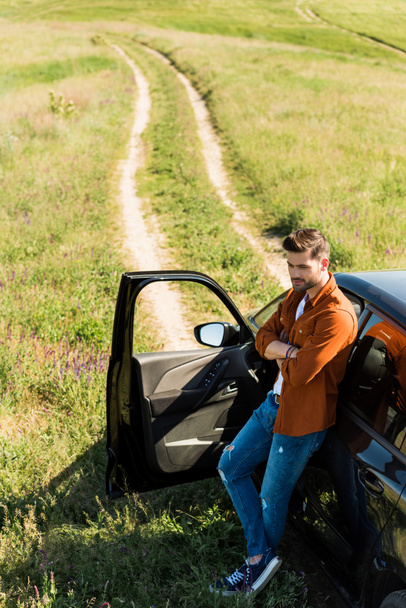 elevated view of young man with crossed hands standing near his car in field  - Foto, afbeelding