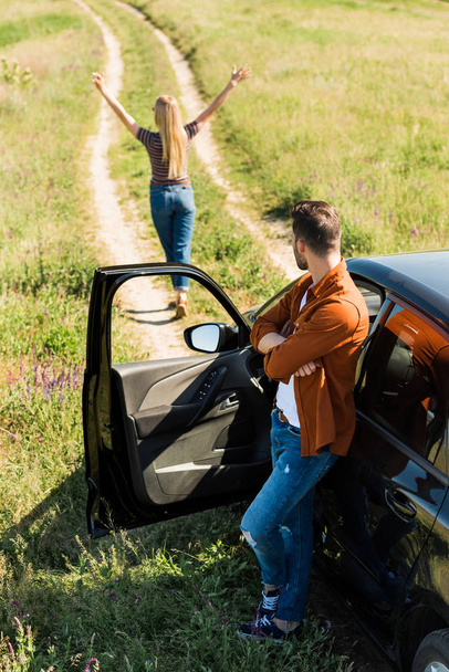 Mann mit gekreuzten Händen steht neben Auto und sieht Freundin mit breiten Armen in ländlichem Gebiet an  - Foto, Bild