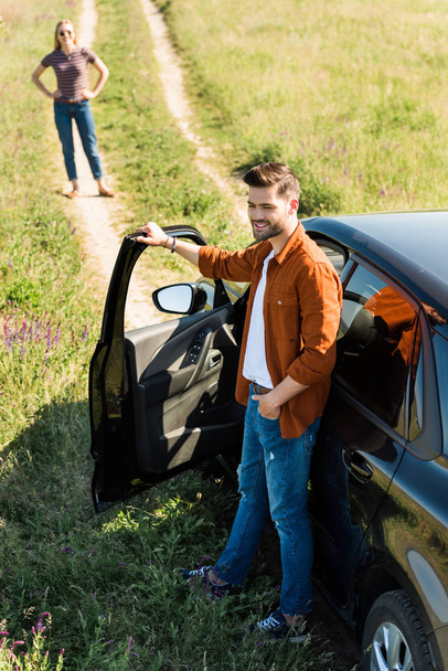 uśmiechnięty mężczyzna młody zamknięcie drzwi samochodu i jego dziewczyna czeka za na wsi łąka  - Zdjęcie, obraz