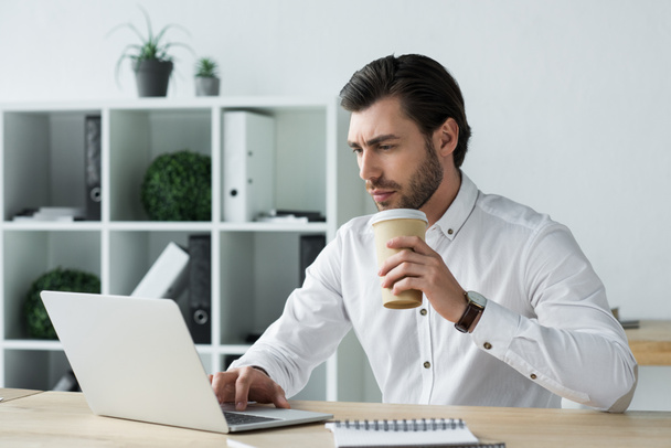 handsome young businessman with paper cup of coffee working with laptop - Fotó, kép