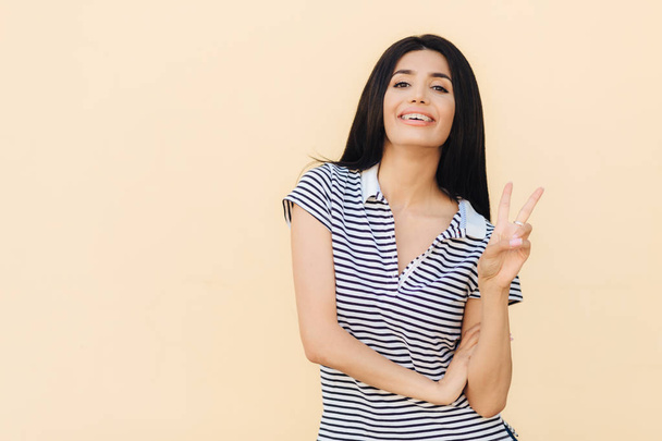 Portrait of cheerful brunette female shows peace gesture, keeps two fingers raised, has pleasant smile, dressed in striped t shirt, poses against studio wall. People and body language concept - Photo, Image