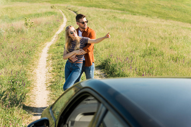 man embracing stylish girlfriend in sunglasses pointing by finger in rural field near car  - 写真・画像