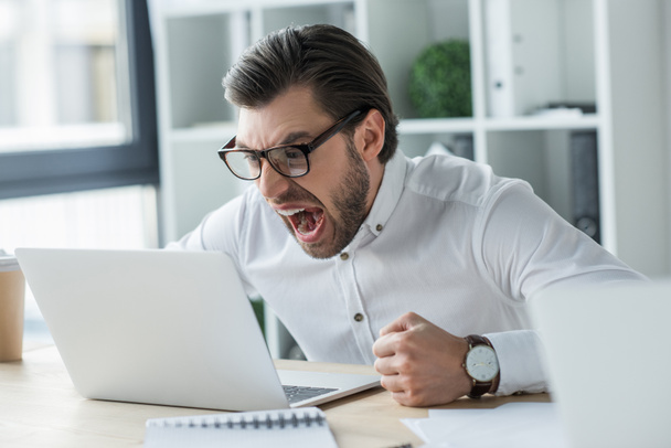 сумасшедший молодой бизнесмен кричит на ноутбук в офисе
 - Фото, изображение