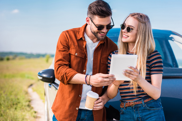 pareja sonriente en gafas de sol de pie con taza de café y tableta digital cerca del coche
  - Foto, imagen