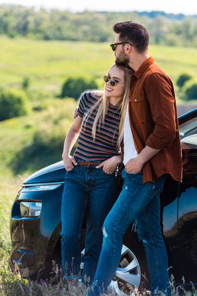 front view of stylish young couple in sunglasses standing near car on rural meadow  - Photo, Image