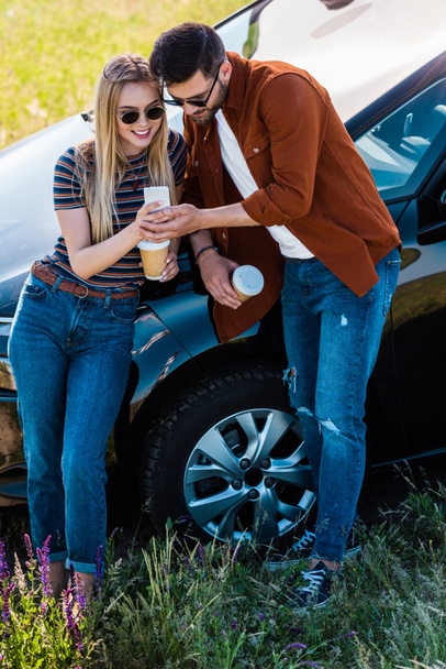 車の近くのスマート フォンの画面を見てコーヒー カップとスタイリッシュなカップルのハイアングル  - 写真・画像