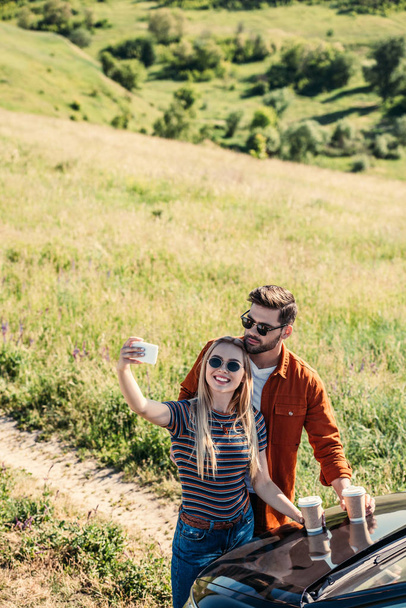high angle view of stylish couple in sunglasses with coffee cups taking selfie on smartphone near car on rural meadow  - Fotografie, Obrázek
