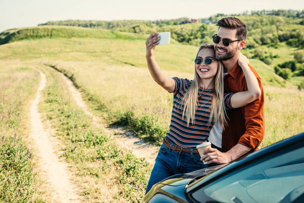 smiling stylish couple in sunglasses with coffee cup taking selfie on smartphone near car on rural meadow  - Фото, зображення