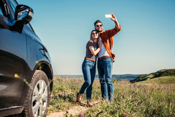 homem elegante tomando selfie com namorada perto de carro no prado rural
  - Foto, Imagem