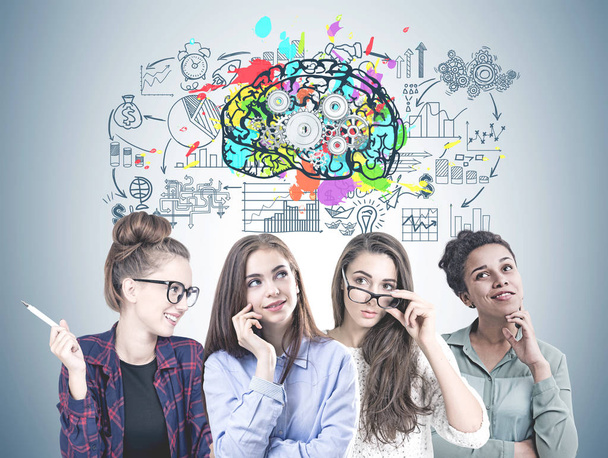 Diverse team of young women wearing glasses and different clothes and hairstyles standing shoulder to shoulder. A gray wall background with a cog brain business sketch. - Photo, Image