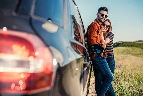 happy stylish couple in sunglasses standing near car on rural meadow  - Photo, Image