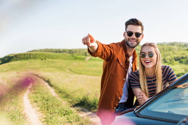 young stylish man pointing by hand to smiling woman near car on rural meadow  - Photo, Image