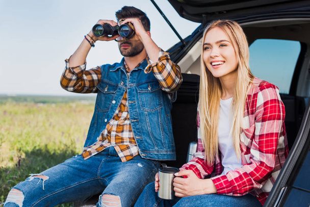 male tourist looking through binoculars while his smiling girlfriend sitting near with coffee cup on car trunk in field - Foto, imagen