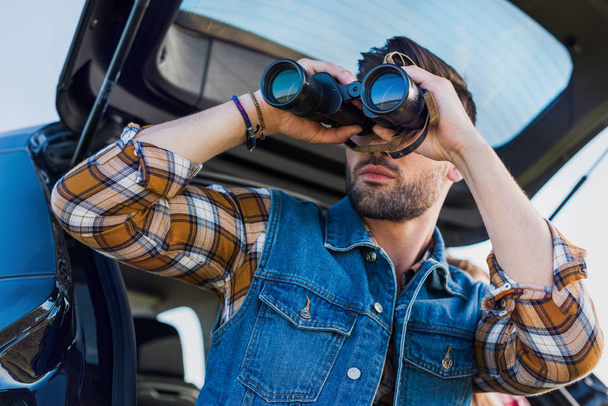 Tiefansicht eines männlichen Touristen, der durch ein Fernglas schaut, während seine Freundin in der Nähe auf dem Kofferraum des Autos sitzt  - Foto, Bild