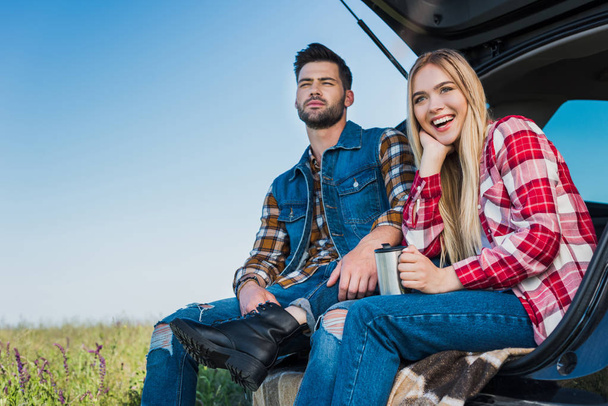 smiling woman with coffee cup and her boyfriend sitting near on car trunk in field - Photo, Image