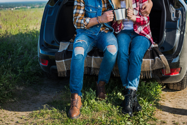 cropped shot of couple of tourists clinking by coffee cups and sitting on car trunk in rural field  - 写真・画像