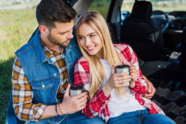 young couple with coffee cups sitting on car trunk in rural field  - Foto, Bild