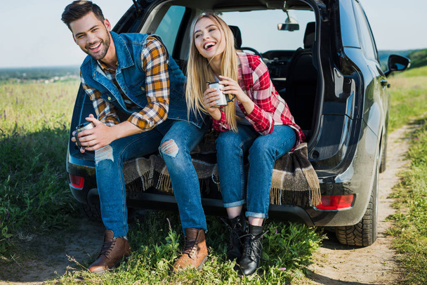 smiling couple of stylish tourists with coffee cups sitting on car trunk in rural field  - Fotó, kép