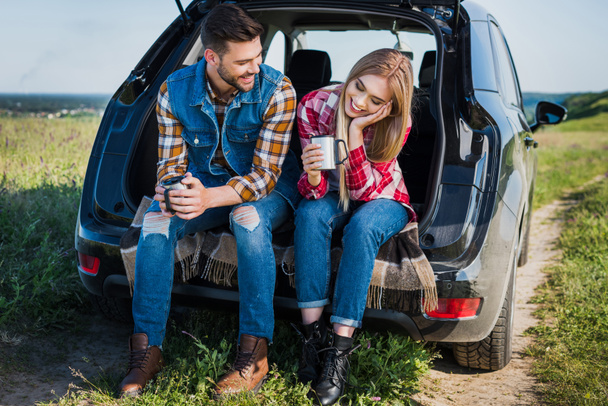 couple avec tasses à café parler entre eux et assis sur le coffre de la voiture dans le domaine rural
  - Photo, image