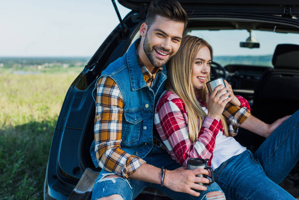 smiling couple with coffee cups sitting on car trunk in rural field  - Φωτογραφία, εικόνα