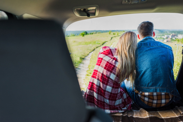 back view of couple of stylish travelers sitting on car trunk in rural field  - Foto, immagini