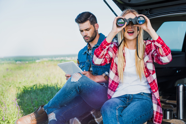 彼氏農村フィールドで車のトランクにデジタル タブレットを使用しながら双眼鏡でみる女性の観光  - 写真・画像