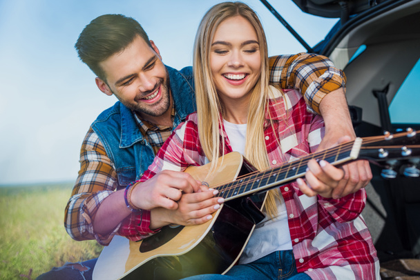 giovane uomo insegnamento sorridente fidanzata a giocare sulla chitarra acustica mentre sono seduti sul bagagliaio auto
  - Foto, immagini