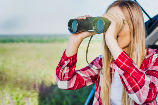 young female traveler looking through binoculars near car in rural field - Photo, Image