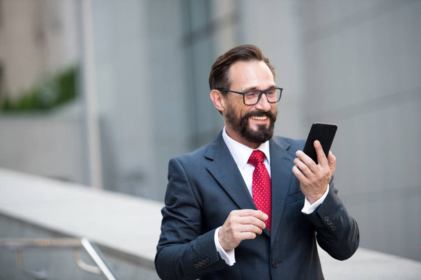 Businessman in glasses using smart phone on office walkway with city building background. Concept of business people using technology. man in suit is searching information on web via mobile phone. - Photo, image