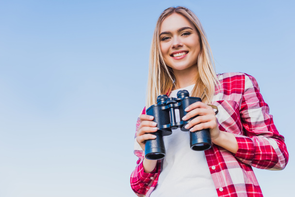 smiling female traveler holding binoculars against blue sky - Photo, Image