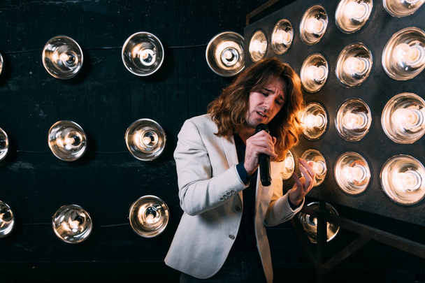 men singer with microphone on the stage, with long hair, against the background of bright lanterns, scene - Photo, image