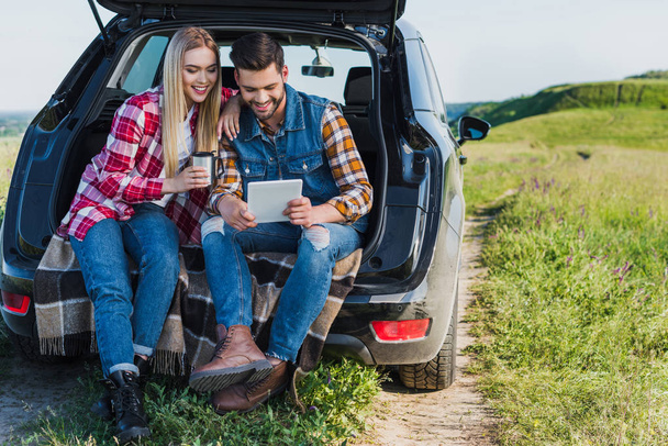 homem feliz usando tablet digital, enquanto sua namorada sentado perto com xícara de café no porta-malas do carro
  - Foto, Imagem